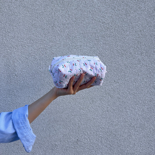Small cherry quilted pouch shown from an angled top view, held in hand against textured wall