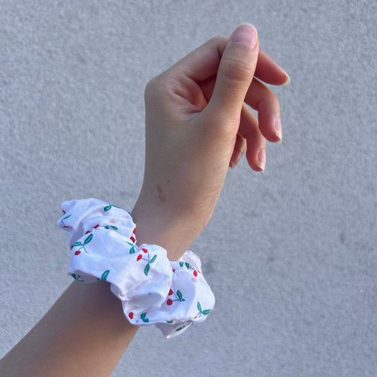 Side view of a cherry-print scrunchie on wrist, hand lifted slightly with fingers curled against a gray background.
