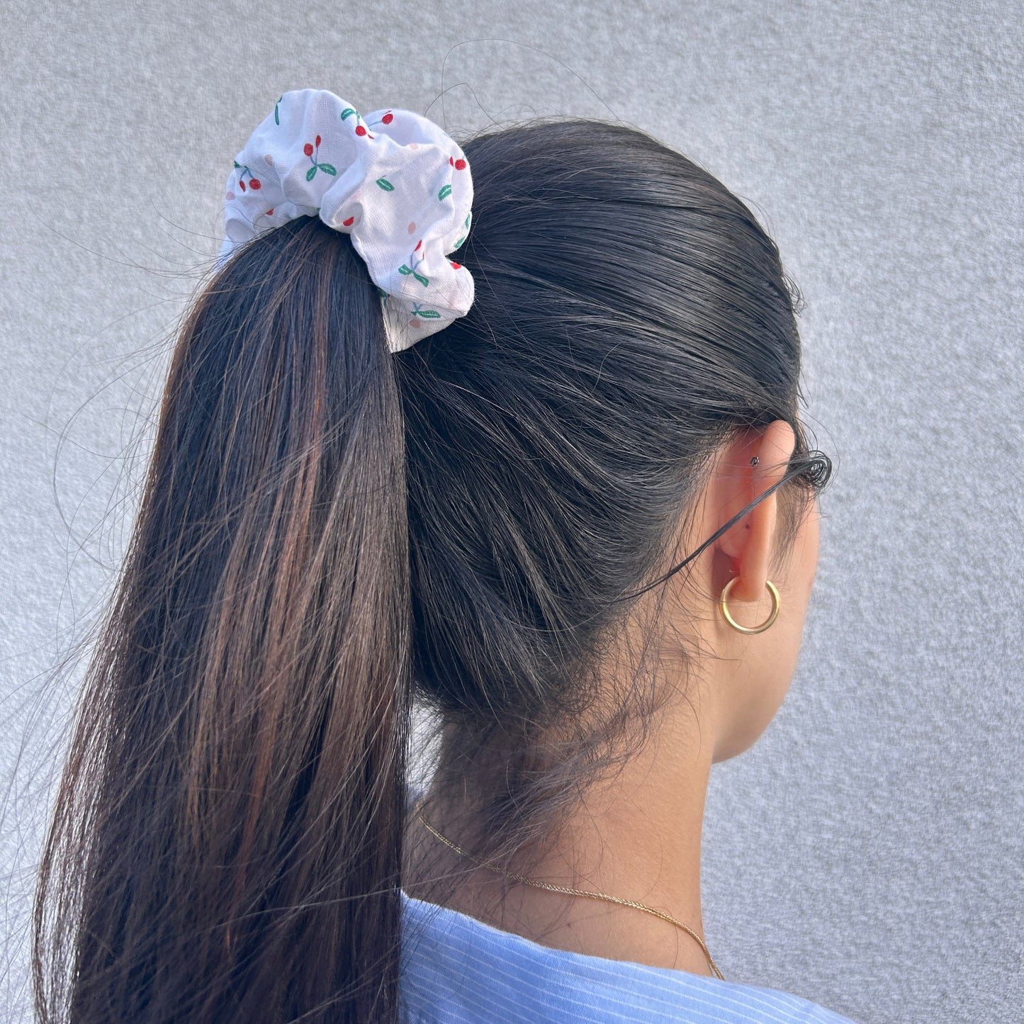 Cherry-patterned scrunchie styled in high ponytail, worn by model with long dark hair, gold hoop earring, and blue collar visible.