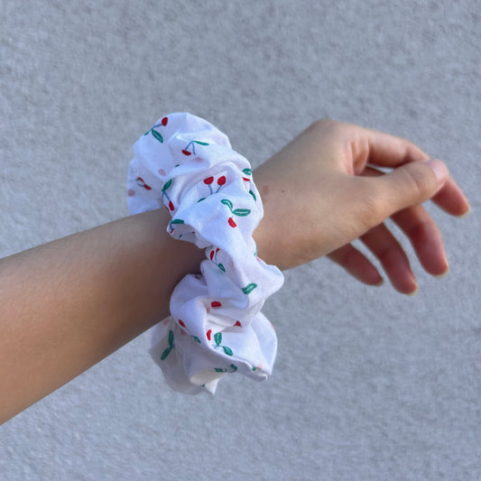 Cherry-patterned scrunchie wrapped around wrist, front view with relaxed arm against a light gray textured wall.